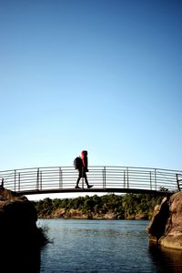 Side view of man walking on footbridge over river against clear sky