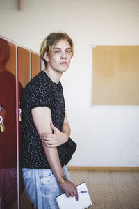 Side view portrait of confident young man holding book leaning on locker at high school