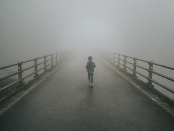 Rear view of boy walking on bridge during foggy weather