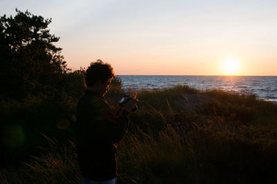 Young boy holding camera while standing on grassy field during sunset