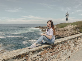 Young woman sitting on retaining wall against sea against sky