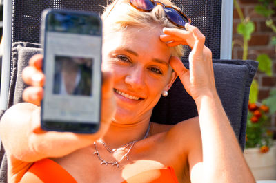Close-up portrait of young woman showing mobile phone while sitting on chair in yard
