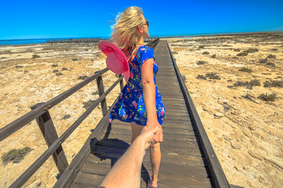 Cropped image of man holding woman hand walking on footbridge against sea and sky