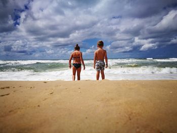 Rear view of siblings standing on beach against cloudy sky