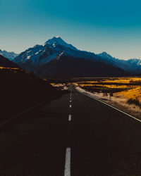 Empty road leading towards a snowcapped mountain peak against a blue sky