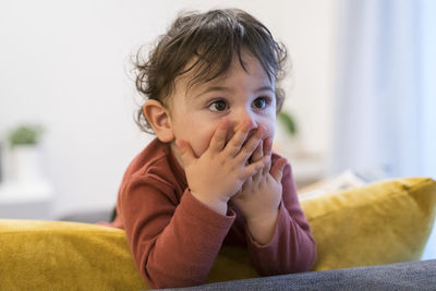 Baby boy covering mouth and looking away while standing on sofa