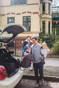 Woman looking at man carrying son while putting luggage in car trunk