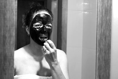 Woman with facial mask applying lipstick while looking in mirror reflection at home