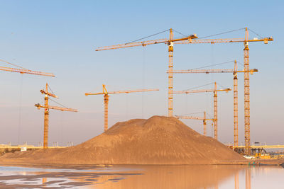 High pile of sand and group high-altitude tower cranes at construction site, sunset sky background.
