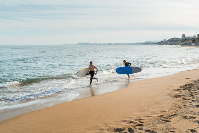 Young man and woman with surfboards in hands running along sandy seashore near waving sea while preparing for surfing together in summer day