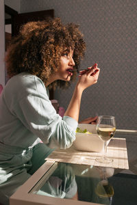 Side view of african american female eating salad while sitting at table with glass of wine and having tasty lunch at home
