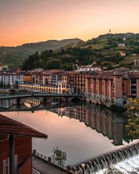 Bridge over river by buildings against sky at sunset