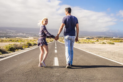 Rear view of young couple holding hands while walking on road against cloudy sky