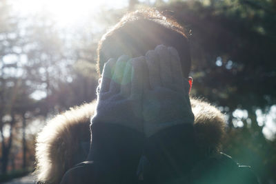 Close-up of man wearing warm clothing covering face with hands during sunny day