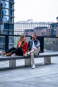 Happy friends sitting on seat against buildings in city