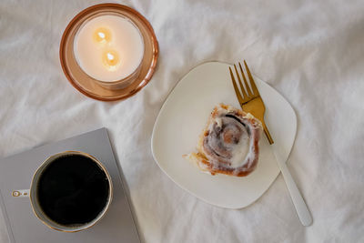 Lifestyle composition with cinnamon bun, coffee and burning candle on the bed.
