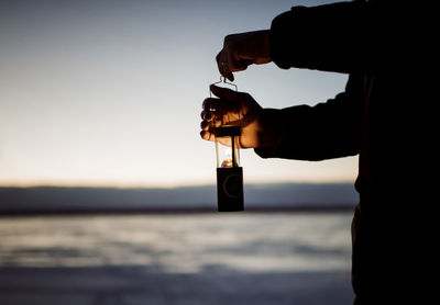 Midsection of silhouette man holding illuminated lantern while standing on snow covered field during sunset