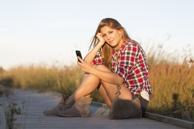 Portrait of smiling young woman using mobile phone against sky