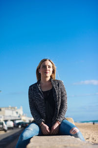 Woman looking away while sitting on wall against blue sky