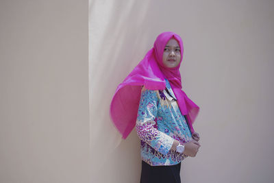 Thoughtful young woman wearing pink hijab looking away while standing against wall