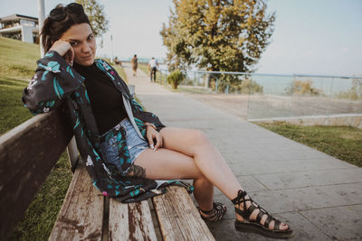 Full length portrait of young woman sitting by footpath on bench