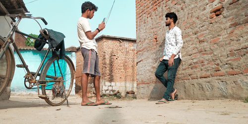 Man photographing male friend standing against brick wall