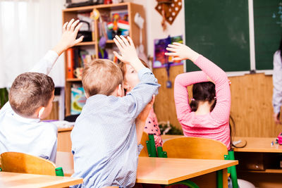 Rear view of kids with arms raised sitting at classroom