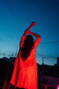 Back view of unrecognizable ethnic female in lingerie looking at camera while resting on terrace under red neon light at night with blue dark sky