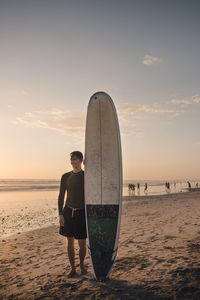 Full length of young man with surfboard standing at beach during sunset