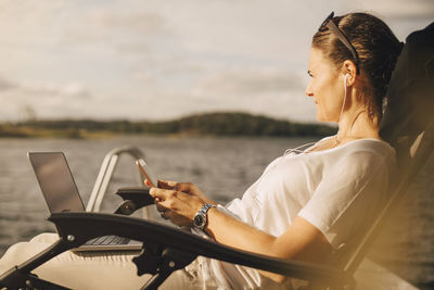 Thoughtful woman with laptop using mobile phone against lake