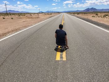 Rear view of man kneeling on empty road against sky