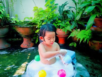 Shirtless girl playing with colorful balls in wading pool against potted plants