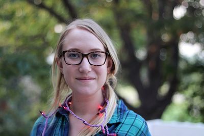 A young woman smiles as she stands outside in purple lights with trees in the background.