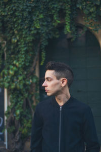 Young man looking away while standing against creeper plants