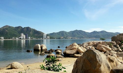 View of rocks by river with buildings in front of mountain range