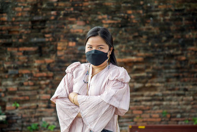 Asian woman in a pink dress crosses her arms wearing a face mask with an old brick wall background.