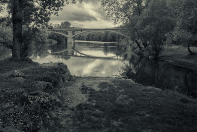 Reflection of arch bridge in river amidst trees against cloudy sky