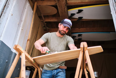 From below crop male worker with hand roller applying glue on plank of longboard on stands in daytime