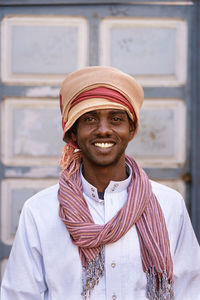 Portrait of content egyptian male wearing traditional headwear looking at camera with toothy smile against blurred background in city