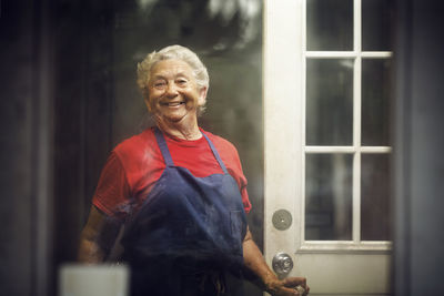 Portrait of smiling owner standing at doorway of kitchen seen through window glass