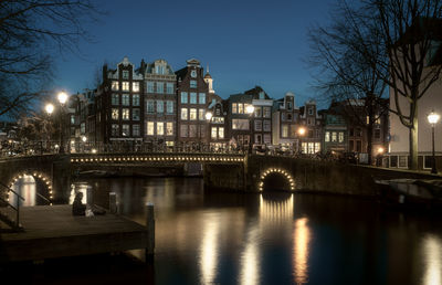 Illuminated bridge over river by buildings against sky at night