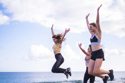 Happy young friends jumping while exercising at beach against sky