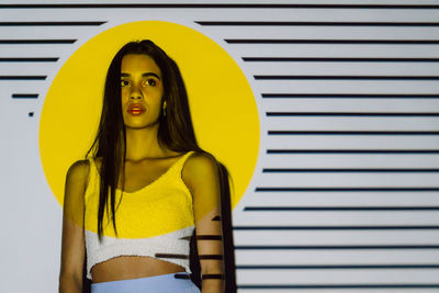 Contemplative young hispanic woman with makeup and long hair looking away in yellow projector light with stripes