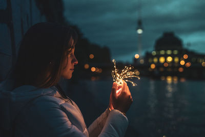 Young woman holding illuminated string light by river at night