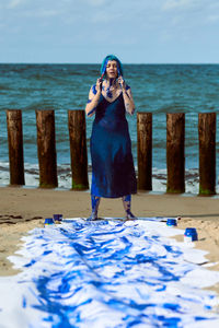 Young woman performance artist in dark blue dress smeared with blue gouache paints dancing on beach