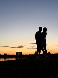 Couple standing at lakeshore against sky during sunset