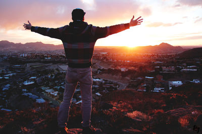 Rear view of man with arms outstretched standing on rock against cityscape against sky during sunset