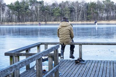 Rear view of man sitting on pier by frozen lake during winter