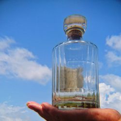 Close-up of hand holding glass bottle against sky