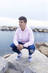 Young man looking away while crouching on rock against sea